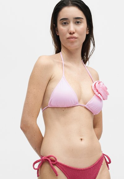 Νεαρή γυναίκα που φοράει ροζ τρίγωνο μπικίνι τοπ με λεπτομέρεια από τριαντάφυλλο και υφασμάτινα σκούρα ροζ μπικίνι κάτω μέρη δεμένα στα πλαινά.