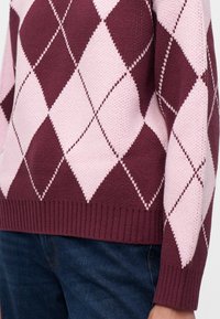 Gros plan d'une personne portant un pull en maille avec un motif losanges rose et bordeaux et un jean en denim bleu.