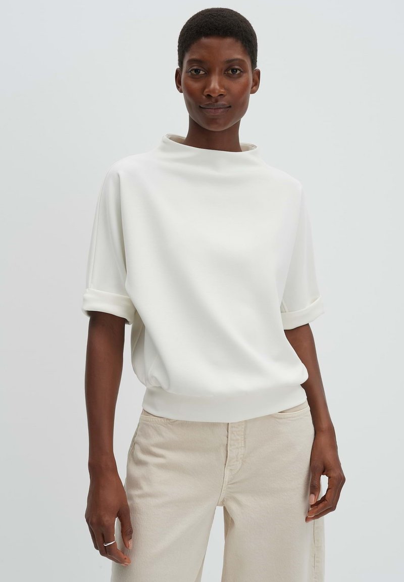 Woman with short hair wearing a white short-sleeve top with rolled cuffs and beige pants, standing against a plain light background.