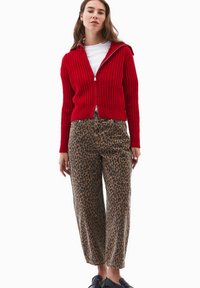 Czerwona dzianinowa bluza na zamek z kapturem, zestawiona z brązowymi spodniami o wysokim stanie w cętki. Modelka stoi w czarnych butach wsuwanych.