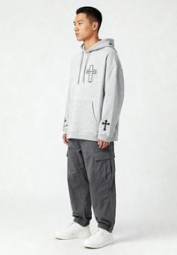 Hoodie grigio con grafiche nere a croce, tasca a marsupio e polsini a coste; abbinato a pantaloni cargo grigi larghi e sneakers bianche.