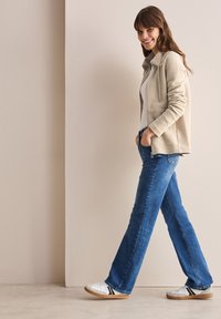 Chaqueta beige texturizada con cremallera, parte superior blanca, jeans acampanados azules y zapatillas blancas con rayas negras, posando frente a un fondo neutro.