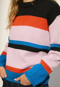 Pull en maille épaisse avec des rayures noires, orange, roses, bleues et noires. Col roulé côtelé et manches larges avec un motif texturé.