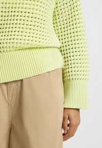 Lichtgroene gebreide trui met een gestructureerd patroon, geribde manchetten en een merketiket aan de zoom, gecombineerd met beige broeken.