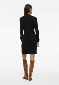 Abito nero a maglia a coste con maniche lunghe, silhouette aderente, lunghezza fino al ginocchio, abbinato a stivali marroni alti fino al ginocchio con tacco e texture liscia.
