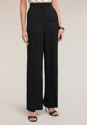 Kobieta w czarnych, szerokich spodniach z wysokim stanem, czarnych sandałkach na obcasie i złotych oraz czarnych bransoletkach na prawej ręce.