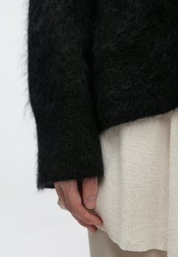 Must hallikas kampsun pikkade varrukatega, tekstureeritud kangas, kombineeritud soonilise helerohelise särgiga. Käsi puhkab äärel, näidates kihte.