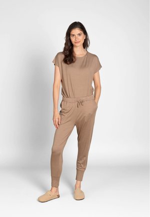 Woman with long dark hair wearing a brown striped jumpsuit and beige slippers standing against a plain light background.