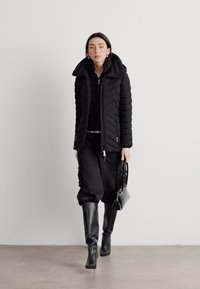 Giacca a vento nera con colletto alto, abbinata a un top nero con zip e pantaloni larghi. Porta una piccola borsa nera a tracolla e indossa stivali fino al ginocchio.
