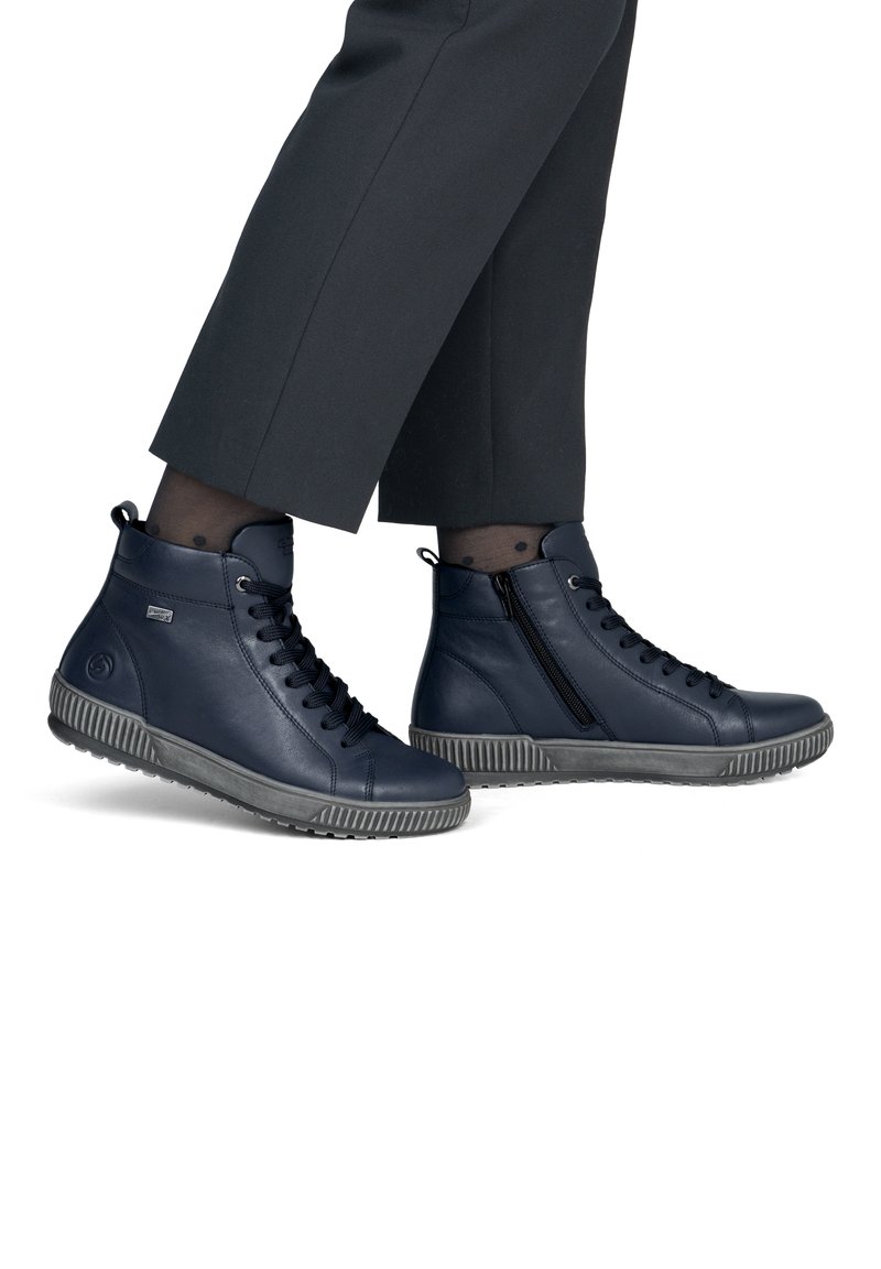 Personne portant un pantalon foncé coupé court et des baskets montantes bleu marine avec des semelles et des lacets noirs, debout sur un fond blanc.