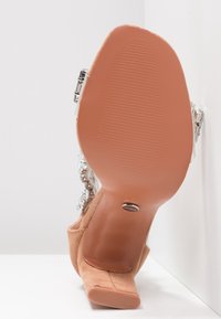 Chaussure à talon haut en daim nude avec semelle en caoutchouc texturée, bride transparente et strass décoratifs sur le côté, présentant un design moderne.