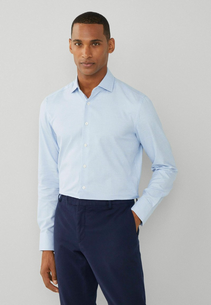 Camicia a scacchi blu chiaro con colletto a punta, maniche lunghe e chiusura con bottoni, abbinata a pantaloni scuri navy. Tessuto dalla superficie liscia.