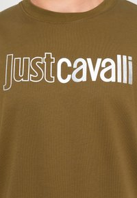 Olivinės spalvos medvilnės marškinėliai su dideliais metaliniais sidabro spalvos užrašais "Just Cavalli" ir klasikiniu apvaliu kaklu.