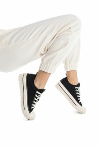 Zapatillas de lona negra con suelas de goma blancas, con punteras redondeadas, diseño de cordones en contraste y suela texturizada.