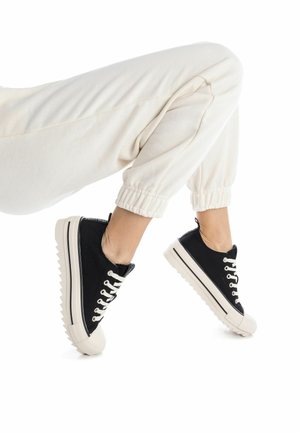 Zapatillas de lona negra con suelas de goma blancas, con punteras redondeadas, diseño de cordones en contraste y suela texturizada.