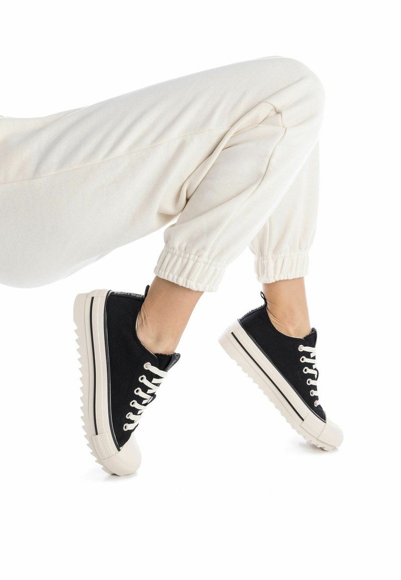 Zapatillas de lona negra con suelas de goma blancas, con punteras redondeadas, diseño de cordones en contraste y suela texturizada.