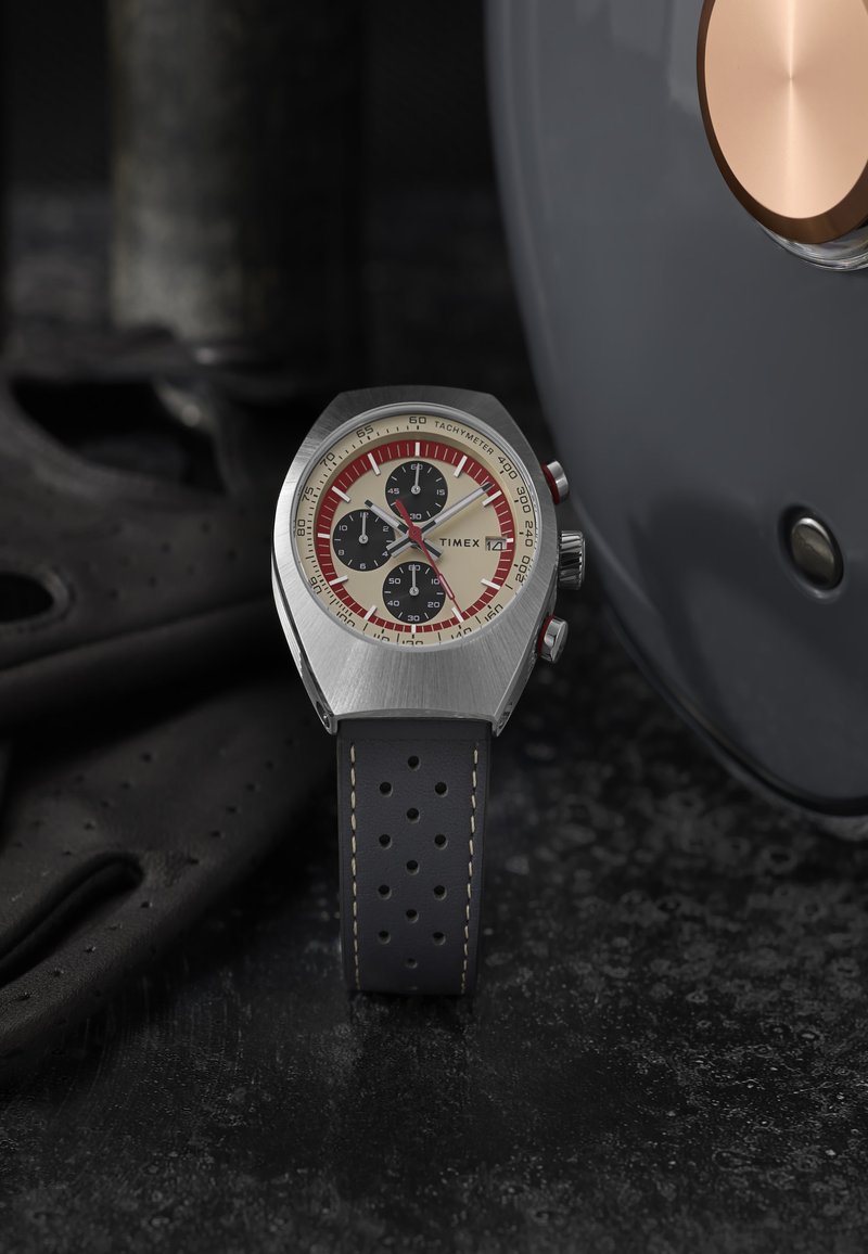 Montre chronographe en acier inoxydable avec un cadran crème, des sous-cadrans noirs, des accents rouges et un bracelet en cuir perforé noir, le tout sur un fond texturé.