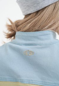 Ljusblå denimjacka med krage och en broderad logo i guld. Mjuk textur med en slät yta. Lätt tyg synligt under kragen.