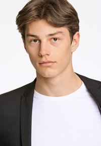 Schwarzer Blazer über einem weißen T-Shirt, Slim Fit Design, glatte Textur, sitzend vor einem schlichten weißen Hintergrund, mit neutralem Ausdruck.