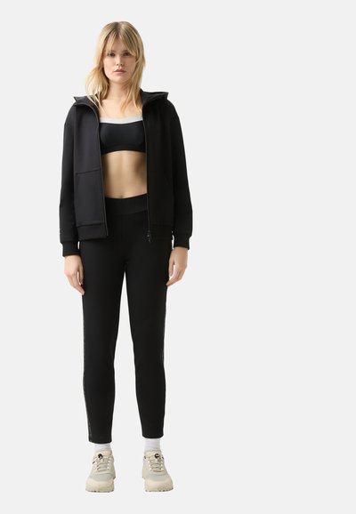 Zwarte zip-up hoodie en sportbeha gecombineerd met zwarte leggings met zijstrepen. Schoenen zijn beige met veters en een gestructureerde zool.