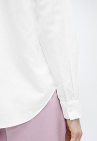 Μανσέτα λευκού πουκάμισου με μακριά μανίκια και κουμπί, φορεμένη πάνω από ανοιχτό ροζ παντελόνι, με έμφαση στις ραφές των πλευρών και τις λεπτομέρειες του μανικιού.