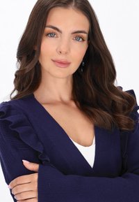 Granatowy kardigan z falbaniastymi ramionami, prążkowaną fakturą i długimi rękawami. Nosi się go na białej bluzce, podkreślając dekolt.