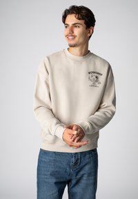 Beige cotton sweatshirt with ribbed cuffs and waistband. Black graphic print on the chest. Paired with blue jeans. Simple, casual design.