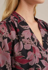 Bluse mit Blumenmuster, die tiefviolette und rosa Blumen auf einem schwarzen Stoff zeigt. V-Ausschnitt mit dezenten Raffung an den Schultern.