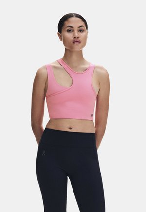 Frau trägt ein pinkfarbenes asymmetrisches Croptop mit Ausschnitten und schwarze hoch taillierte Leggings, steht mit den Händen hinter dem Rücken.