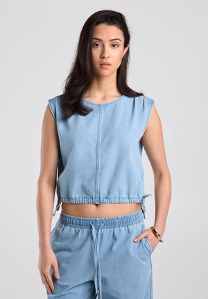 Jeune femme aux longs cheveux foncés, portant un crop top en jean bleu clair sans manches et un pantalon assorti à taille élastique, debout avec une main dans la poche.