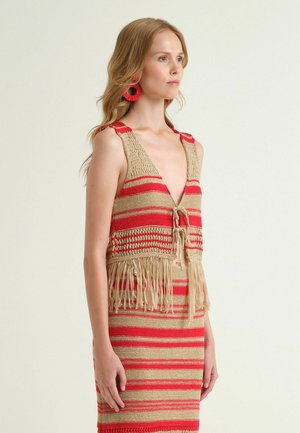 Femme portant un gilet en crochet beige et rouge à rayures avec des franges, assorti à une jupe, accessoirisé de grandes boucles d'oreilles rouges, sur un fond uni.