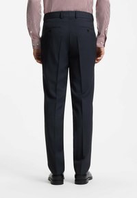 Homme portant un pantalon formel bleu marine, des chaussures habillées noires et une chemise à manches longues rayée rouge et blanche, debout de dos sur fond blanc.
