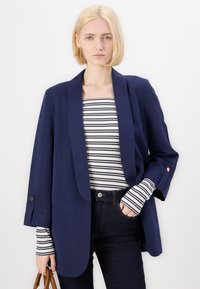 Marinblå blazer med sjalkrage, lätt material och knappade ärmar, i kombination med en randig långärmad topp och mörka jeans.