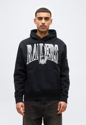 Mladý muž v čiernej mikine Raiders s predným vreckom a logom, stojaci pred jednoduchým svetlým pozadím.