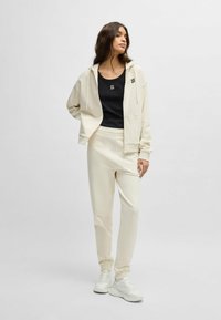 Felpa con zip color avorio e coulisse, con logo nero sul petto; abbinata a pantaloni jogger coordinati. Sneakers bianche completano l'outfit. Texture liscia.