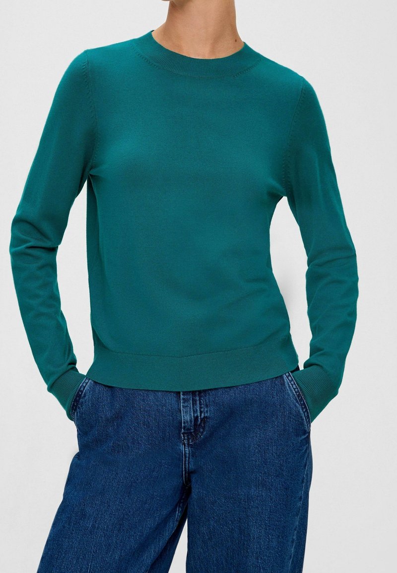 Suéter teal con cuello redondo, mangas largas y puños acanalados, combinado con jeans de mezclilla azul oscuro. Textura suave y ajuste clásico.
