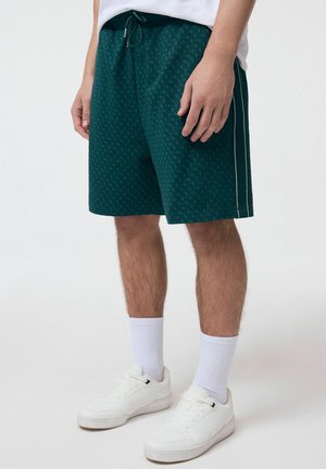 Mężczyzna ubrany w ciemnozielone szorty w wzory z bocznymi paskami, białe skarpetki i białe trampki, stojący na jednolitym tle.