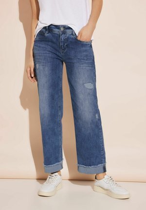 Personne portant un jean bleu coupe décontractée avec des revers roulés et des baskets blanches, debout devant un fond beige.