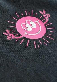 Svart tyg med en stor rosa leendesol som har höjda armar och ett fredstecken, detaljfyld med vita och rosa konturer samt solstråle-accenter.