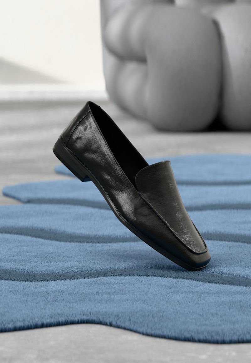 Chaussure en cuir noir à enfiler avec une texture lisse, bout pointu et semelle plate basse, placée au-dessus d'un tapis bleu à motifs.
