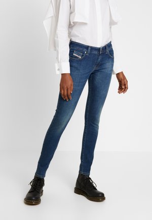 Personne portant un jean skinny bleu, des bottes noires à lacets et une chemise blanche à manches longues avec un vêtement blanc drapé sur les épaules.