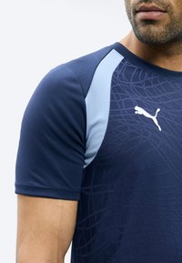 Άνδρας που φοράει μπλε σκούρο κοντομάνικο μπλουζάκι Puma με ανοιχτό μπλε πάνελς στους ώμους και διακριτικό γεωμετρικό σχέδιο μπροστά.