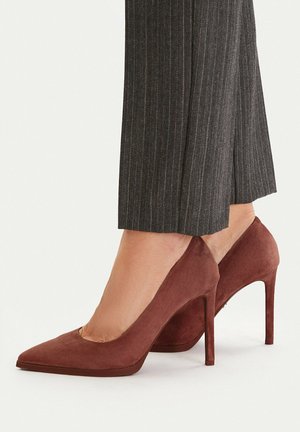 Brązowe szpilki z zamszu o spiczastym nosku noszone z ciemnoszarymi, prążkowanymi, skróconymi spodniami, widoczne od kostki do podłogi na jasnym tle.