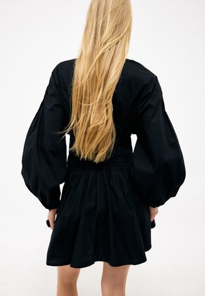 Ženska z dolgimi blond lasmi, oblečena v črno obleko z volumniziranimi dolgimi rokavi, stoji pred enobarvnim belim ozadjem.