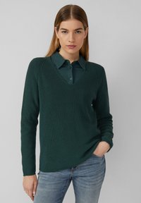 Dunkelgrüner gestrickter Pullover mit V-Ausschnitt, gerippter Textur und langen Ärmeln, über ein kragenloses Hemd getragen, kombiniert mit blauen Jeans.