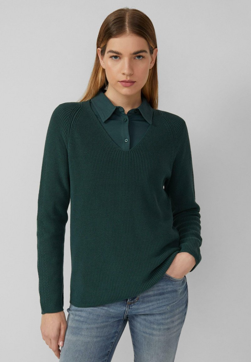 Dunkelgrüner gestrickter Pullover mit V-Ausschnitt, gerippter Textur und langen Ärmeln, über ein kragenloses Hemd getragen, kombiniert mit blauen Jeans.