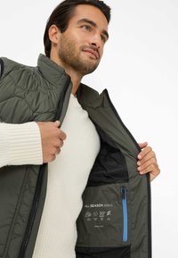 Gestepptes olivgrünes Weste mit Reißverschluss vorne, schwarzem Innenfutter und einer blauen Tasche; enthält Pflegehinweise, die im Inneren gedruckt sind.