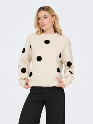 Donna con capelli biondi che indossa un maglione color crema con grandi pois neri e pantaloni neri, mani che aggiustano l'orlo del maglione.