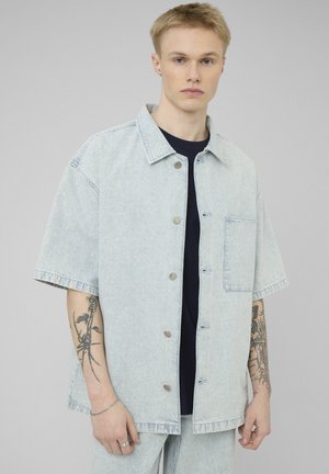 Jeune personne aux cheveux blonds courts portant une chemise à manches courtes rayée bleu clair sur un t-shirt noir, montrant des tatouages sur les deux avant-bras.