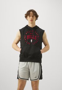 Črna brezrokavna kapuca z rdečim grafičnim napisom "BULLS" in kenguru žepom, v kombinaciji s sivimi in črnimi športnimi kratkimi hlačami z bočnimi črtami.